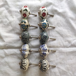 Puede incluir: Conjunto de diez pomos de armario de cerámica con varios patrones florales y geométricos en tonos de azul, gris, blanco y rojo. Los pomos tienen una base de metal plateado y un tornillo.