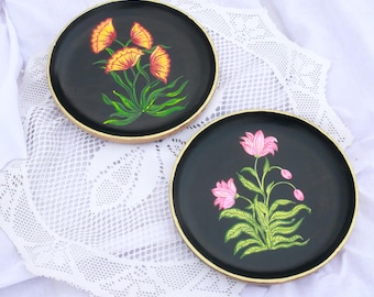 May include: Two round black trays with gold rims, each featuring a hand-painted floral design. One tray has yellow flowers with green leaves, while the other has pink flowers with green leaves.