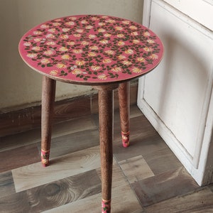 May include: A round wooden side table with a pink top featuring a floral pattern in white and yellow. The table has three wooden legs with decorative painted details at the base. The table is approximately 18 inches in diameter.