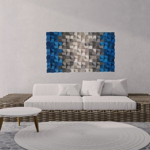 Puede incluir: Un sofá blanco con una base de mimbre tejido se encuentra en una sala de estar moderna con una gran obra de arte de pared geométrica azul y gris encima. Una pequeña mesa de café blanca se encuentra frente al sofá y una planta de cactus en maceta se encuentra a la derecha de la imagen.