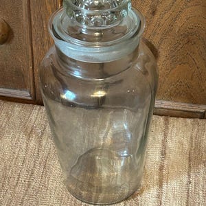May include: A clear glass apothecary jar with a decorative, ridged lid. The jar is cylindrical with a wide mouth and a clear glass lid. The jar is sitting on a textured, tan surface.