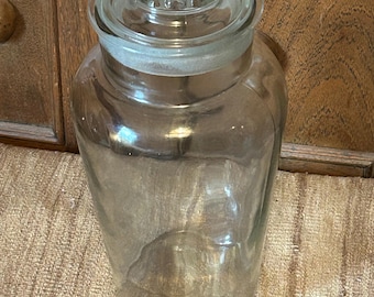 May include: A clear glass apothecary jar with a decorative, ridged lid. The jar is cylindrical with a wide mouth and a clear glass lid. The jar is sitting on a textured, tan surface.