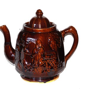 May include: A brown ceramic teapot with a lid and a handle. The teapot is decorated with a raised design of people and trees.