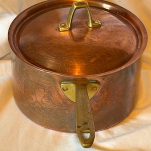 May include: A round copper pot with a matching lid and a brass handle. The pot features a polished copper exterior with visible surface variations and a sturdy brass handle attached with a triangular plate. The lid has a central brass handle.