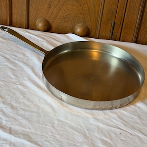May include: A round, shallow metal frying pan with a long, slender handle extending from one side. The pan features a polished, reflective interior surface and a metallic exterior with visible signs of wear and minor discoloration along the base.