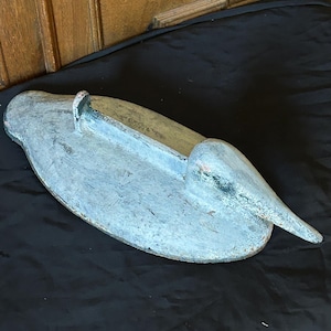 May include: A weathered, hand-carved wooden pelican sculpture, painted in shades of blue and gray. The bird has a long beak and is resting on a black surface. The sculpture is approximately 18 inches long.