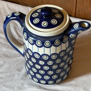 May include: A ceramic teapot with a lid, handle, and spout. The teapot is primarily blue with a white band and a pattern of blue circles with red and green centers. The lid has a matching design.