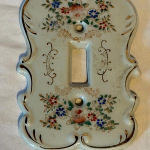 May include: A light-colored ceramic light switch plate with a scalloped edge and gold trim. The surface features hand-painted floral motifs in red, blue, and green, with symmetrical scrollwork patterns flanking the central rectangular toggle opening.