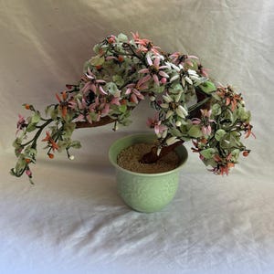 May include: A decorative bonsai tree sculpture featuring light green jade-like leaves and delicate pink and white flower petals attached to dark brown branches. The tree is planted in a pale green ceramic pot with a subtle embossed floral pattern.