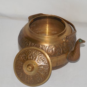 May include: A brass teapot with an ornate floral design and a green glass accent on the spout. The teapot has a matching lid with a floral design.