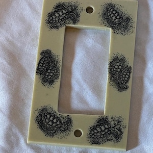May include: Off-white light switch cover featuring six black and white sea turtle illustrations. The rectangular cover has a single rectangular opening and two circular holes for mounting. The sea turtle designs are detailed and surrounded by dotted shading.