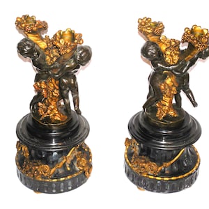 May include: A pair of ornate gold and black candle holders featuring cherubs holding up a grapevine with three candle cups. The base is made of black marble with gold accents.