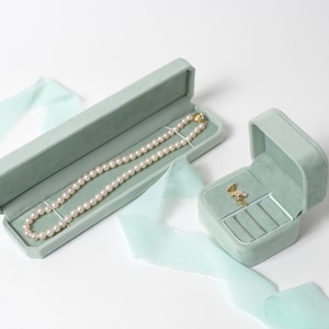 May include: Two light green velvet jewelry boxes. The larger box contains a pearl necklace. The smaller box has a ring holder and a small gold pendant inside.