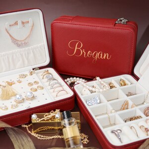 以下が含まれることがあります： 蓋に「Brogan」という名前が刻まれた赤い革製のジュエリーボックス。リング、イヤリング、ネックレス、ブレスレットを収納できる複数のコンパートメントがあります。