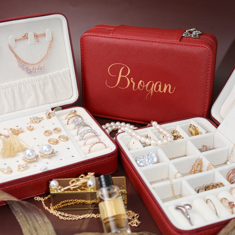 以下が含まれることがあります： 蓋に「Brogan」という名前が刻まれた赤い革製のジュエリーボックス。リング、イヤリング、ネックレス、ブレスレットを収納できる複数のコンパートメントがあります。