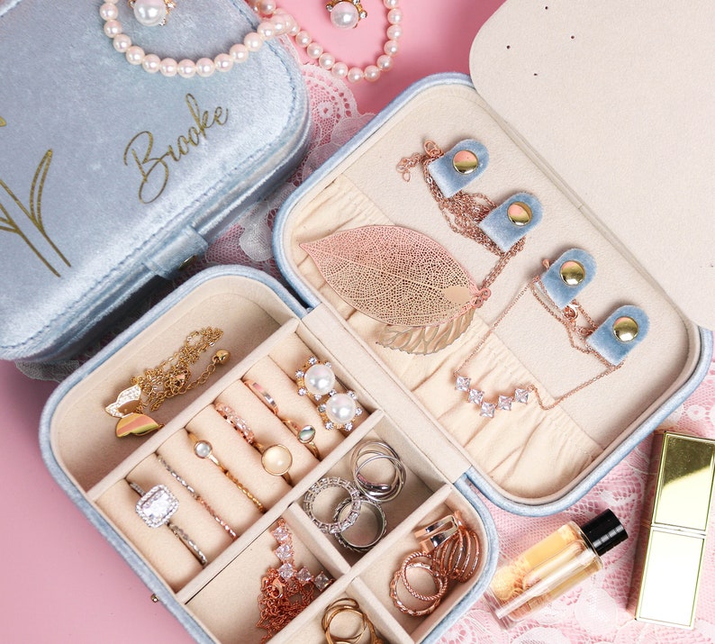 May include: A light blue velvet jewelry box with the name "Brooke" on the lid. The box has multiple compartments for storing rings, earrings, and necklaces. The box is open and shows a variety of jewelry inside.