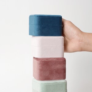 May include: Stack of four velvet jewelry boxes in different colors: teal, light pink, maroon, and light green. The boxes are stacked on top of each other.