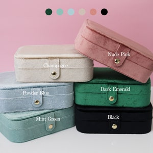 May include: Six velvet jewelry boxes in different colors, stacked on top of each other. The colors are: Champagne, Powder Blue, Mint Green, Dark Emerald, Nude Pink, and Black. Each box has a gold button closure and the color name is printed on the front.