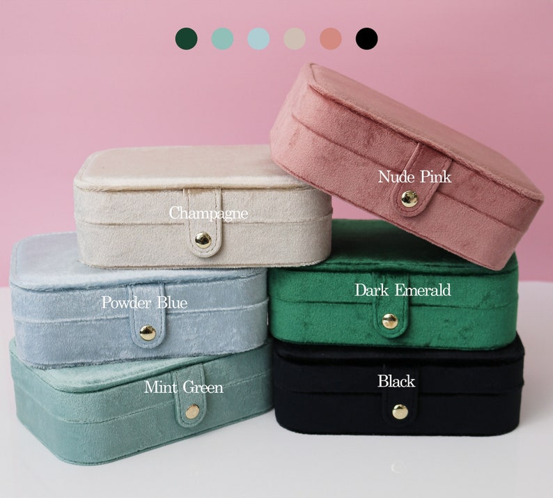 May include: Six velvet jewelry boxes in different colors, stacked on top of each other. The colors are: Champagne, Powder Blue, Mint Green, Dark Emerald, Nude Pink, and Black. Each box has a gold button closure and the color name is printed on the front.