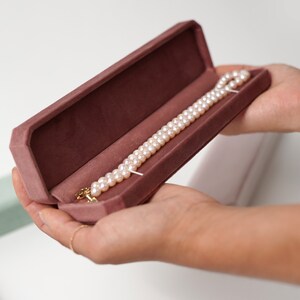 May include: A pearl necklace in a pink velvet jewelry box. The necklace is made of white pearls and has a gold clasp.