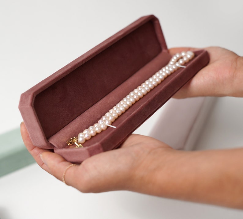 May include: A pearl necklace in a pink velvet jewelry box. The necklace is made of white pearls and has a gold clasp.