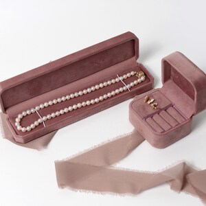 May include: Two velvet jewelry boxes, one long and one square, both in a dusty rose color. The long box contains a pearl necklace, and the square box contains a pair of earrings and rings.