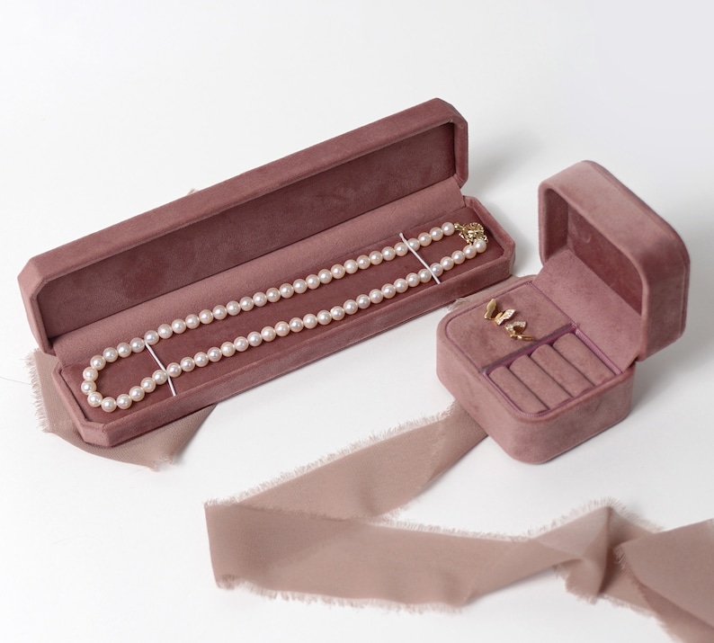 May include: Two velvet jewelry boxes, one long and one square, both in a dusty rose color. The long box contains a pearl necklace, and the square box contains a pair of earrings and rings.