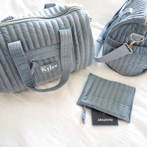 May include: A set of two gray quilted travel bags with gold hardware. The larger bag has a zipper closure and a name tag that reads "Kylee". The smaller bag has a zipper closure and is designed to hold a passport.
