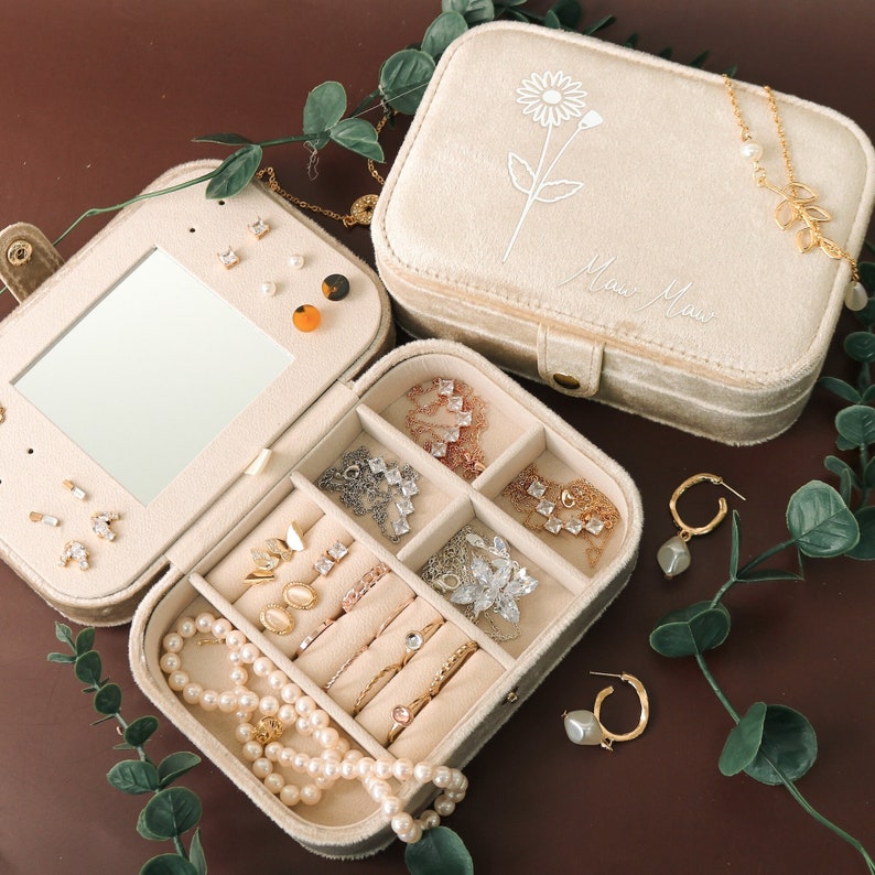 May include: A beige velvet jewelry box with a mirror and compartments for storing necklaces, earrings, and rings. The box has a floral design and the words "Mom Mom" on the lid.