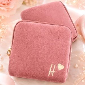 May include: A square, dusty rose velvet jewelry case featuring a gold-colored zipper closure. The front of the case is embroidered with a gold-colored letter H followed by a small gold-colored heart symbol. The case has a soft, textured finish.