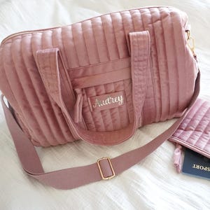 May include: A pink quilted travel bag with a long adjustable strap and a smaller matching pouch. The bag has a zipper closure and a front pocket with the name "Audrey" embroidered on it.