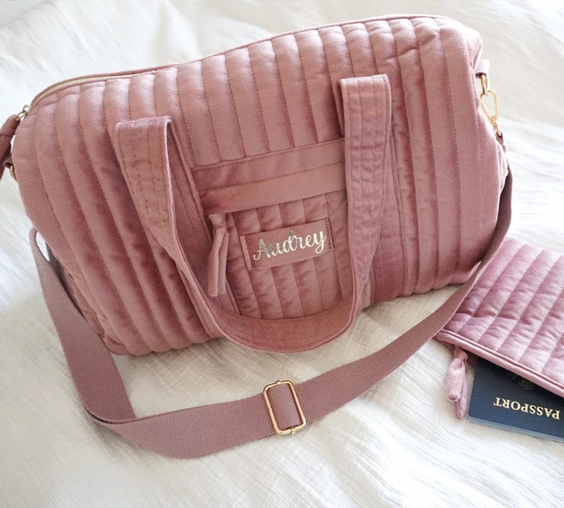 May include: A pink quilted travel bag with a long adjustable strap and a smaller matching pouch. The bag has a zipper closure and a front pocket with the name "Audrey" embroidered on it.