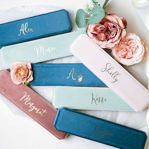 May include: A set of ten personalized velvet jewelry boxes in various colors, including pink, blue, and green. Each box has a gold inscription with a different name, such as Alex, Maria, Amy, Shelly, and Kerr.