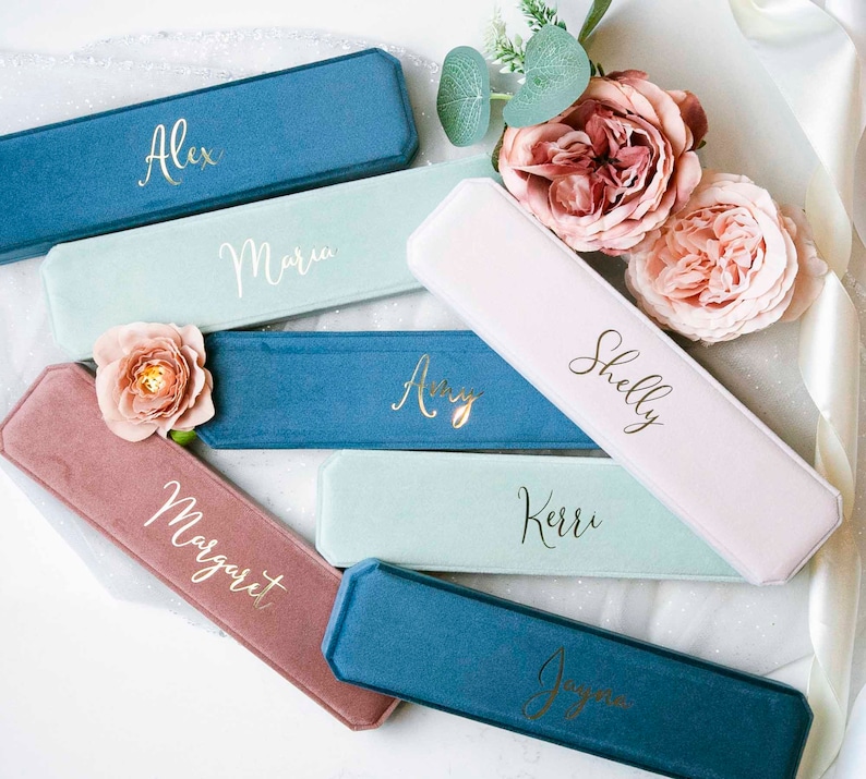 May include: A set of ten personalized velvet jewelry boxes in various colors, including pink, blue, and green. Each box has a gold inscription with a different name, such as Alex, Maria, Amy, Shelly, and Kerr.