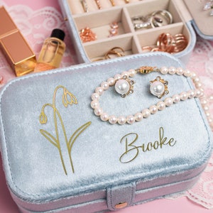 May include: A light blue velvet jewelry box with a gold floral design and the name "Brooke" inscribed on the lid. The box is open and contains a pearl necklace and a pair of pearl earrings.