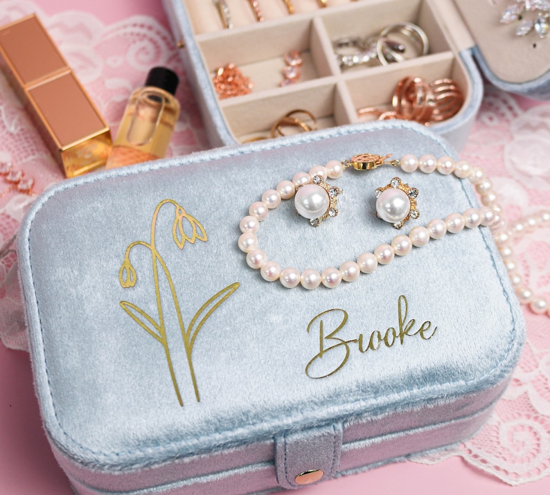 May include: A light blue velvet jewelry box with a gold floral design and the name "Brooke" inscribed on the lid. The box is open and contains a pearl necklace and a pair of pearl earrings.