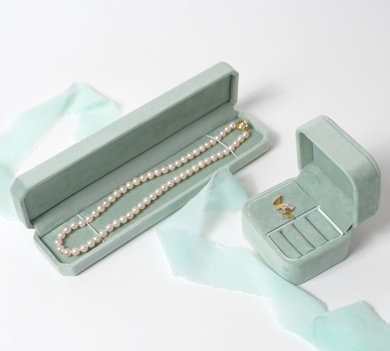 May include: Two light green velvet jewelry boxes. The larger box contains a pearl necklace. The smaller box has a ring holder and a small gold pendant inside.