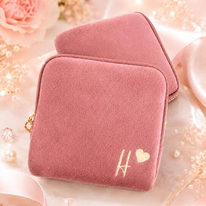May include: A square, dusty rose velvet jewelry case featuring a gold-colored zipper closure. The front of the case is embellished with a gold-colored letter H and a small heart icon. The case has a soft, textured surface and a structured shape.