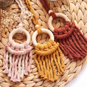 May include: Three macrame wall hangings with wooden rings in shades of pink, mustard yellow, and rust. The handmade pieces feature intricate knotting and fringe details, ideal for adding a touch of bohemian style to any space. The background is a woven mat.