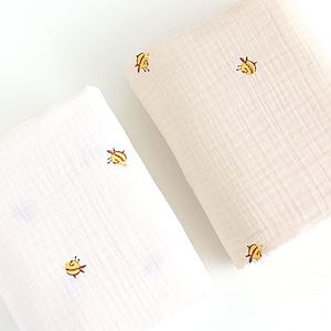 Peut inclure: Deux langes en mousseline blanche avec une texture subtile et un petit motif d'abeille en jaune et brun. Les langes sont pliés et disposés sur une surface blanche.