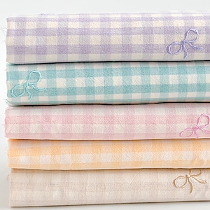 May include: A pile of five folded fabrics in pastel shades: lilac, turquoise, rose, peach, and beige. Each fabric features a gingham check and an embroidered bow. The fabrics are neatly stacked, with the bow visible on each layer.
