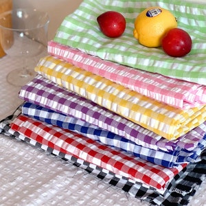 May include: A stack of eight gingham fabric napkins in various colours, including green, pink, yellow, purple, blue, red, and black. The napkins are folded and stacked on a white textured surface.