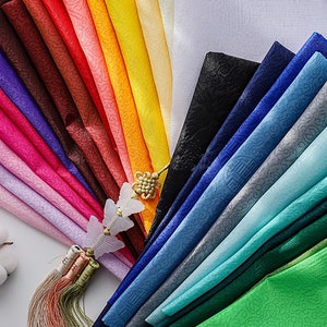 Op de afbeelding: Een verzameling kleurrijke stofstalen in verschillende tinten blauw, groen, roze, rood, oranje, geel en wit. De stof heeft een subtiel patroon en is in een waaier vorm gerangschikt.