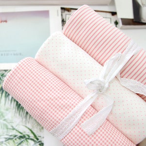 Puede incluir: Cuatro rollos de tela con estampados rosas y blancos, incluyendo rayas, lunares y cuadros vichy. Los rollos están atados con cinta de encaje blanco. Las telas son probablemente para proyectos de costura o manualidades.