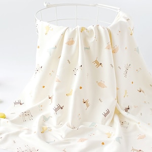 May include: A white muslin baby blanket with a pattern of small, colorful animals. The blanket is folded and draped over a white metal basket.