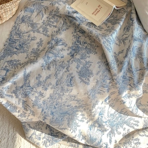 Pode incluir: Uma manta branca com um padrão de toile azul que apresenta um desenho repetitivo de árvores, flores e pássaros. A manta está dobrada e drapeada sobre um sofá branco.