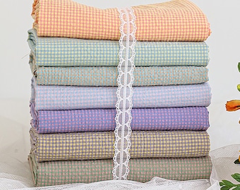 May include: A stack of eight fabric squares in various pastel colors with a gingham pattern. The fabric squares are arranged in a stack with a white lace ribbon tied around the middle.