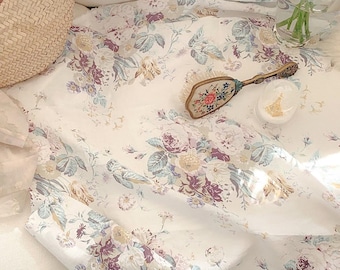 May include: A white fabric with a floral pattern in shades of purple, blue, and yellow. The fabric is folded and draped over a white surface. There is a small, white, round container and a vintage hand mirror with floral embroidery on the fabric.