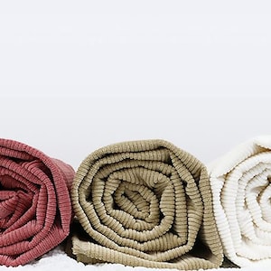 Op de afbeelding: Drie opgerolde dekens in verschillende kleuren: een diep rood, een lichtbruin en een wit. De dekens zijn gemaakt van een zachte, textuurrijke stof.