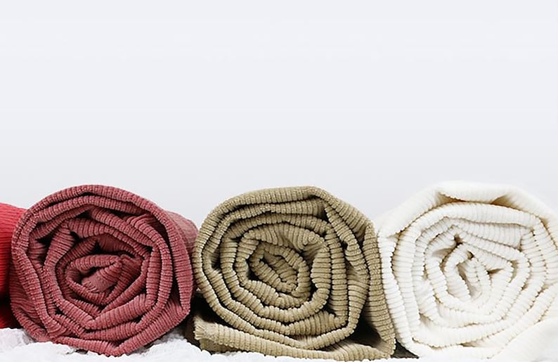 Op de afbeelding: Drie opgerolde dekens in verschillende kleuren: een diep rood, een lichtbruin en een wit. De dekens zijn gemaakt van een zachte, textuurrijke stof.